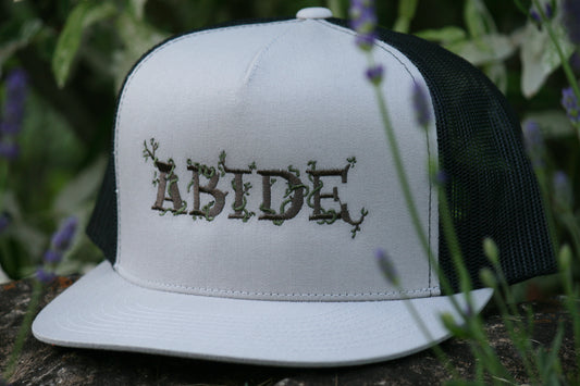 Silver foam trucker hat with black mesh in the back. It has the word abide in the middle of the forehead embroidered. The letters are brown and have moss weaving between the letters. The hat is outside on a rock surrounded by purple flowers.