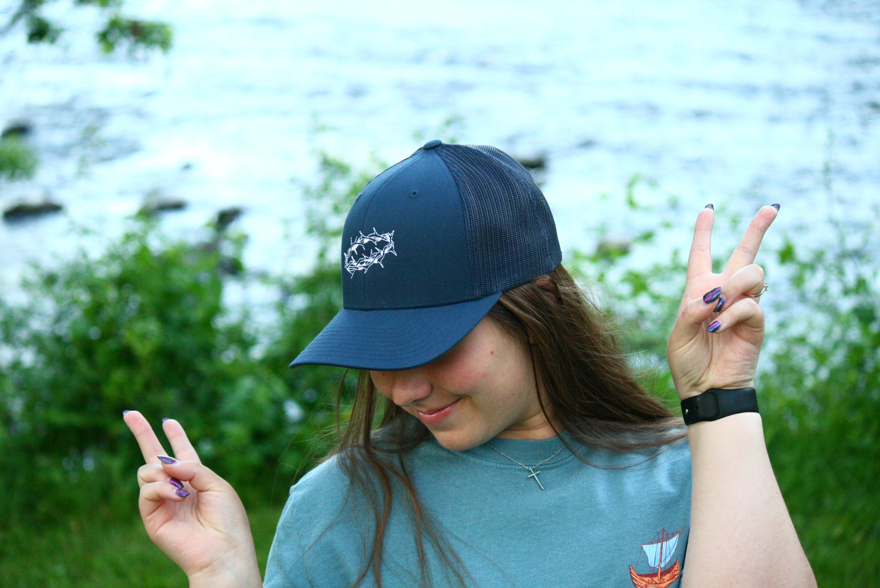 Girl wearing navy blue hat with hands making peace sign. Hat has a white crown of thorns embroidered on top. Surrounded by bushes.