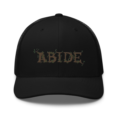 Black trucker hat with the letters Abide on the front. The letters are in brown with green vines wrapped around. The Abide is embroidered on the hat.