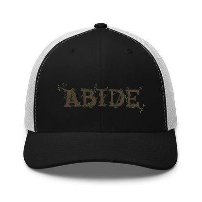 Black trucker hat with white mesh with the letters Abide on the front. The letters are in brown with green vines wrapped around. The Abide is embroidered on the hat.