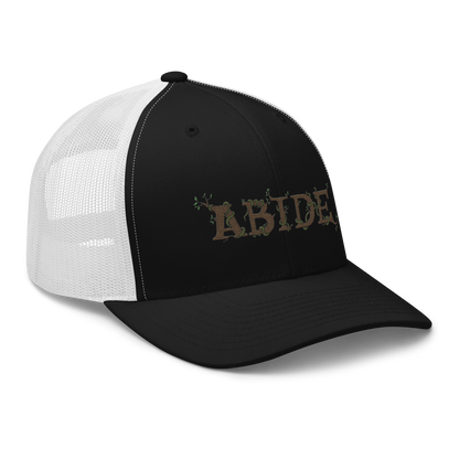 Black trucker hat with white mesh with the letters Abide on the front. The letters are in brown with green vines wrapped around. The Abide is embroidered on the hat.