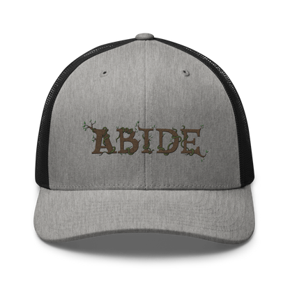 Grey trucker hat with black mesh with the letters Abide on the front. The letters are in brown with green vines wrapped around. The Abide is embroidered on the hat.