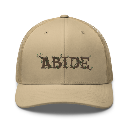 Desert tan trucker hat with the letters Abide on the front. The letters are in brown with green vines wrapped around. The Abide is embroidered on the hat.