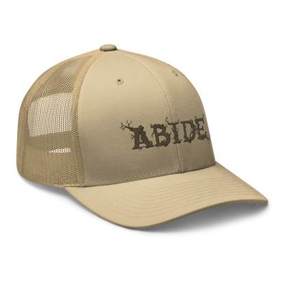 Desert tan trucker hat with the letters Abide on the front. The letters are in brown with green vines wrapped around. The Abide is embroidered on the hat.