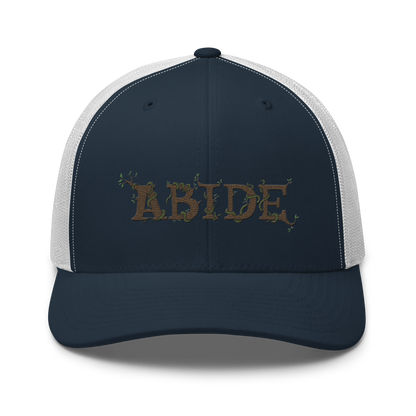 Navy blue trucker hat with white mesh with the letters Abide on the front. The letters are in brown with green vines wrapped around. The Abide is embroidered on the hat.