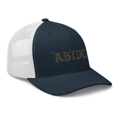 Navy blue trucker hat with white mesh with the letters Abide on the front. The letters are in brown with green vines wrapped around. The Abide is embroidered on the hat.