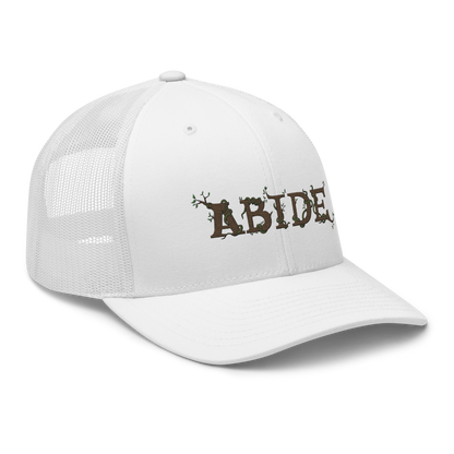 White trucker hat with the letters Abide on the front. The letters are in brown with green vines wrapped around. The Abide is embroidered on the hat.